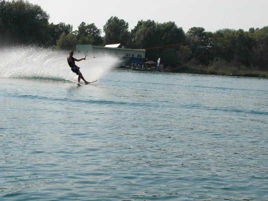 Wasserski auf dem Kiessee Eilenburg Person fährt elegant Wasserski über funkelndes Wasser, im Hintergrund dichter Wald und Gebäudestrukturen.