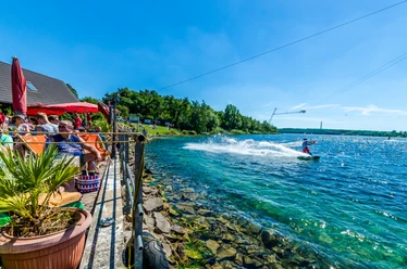 Osoba jeździ na wakeboardzie po krystalicznie czystej wodzie jeziora Kulkwitz, podczas gdy goście relaksują się na słonecznym tarasie.
