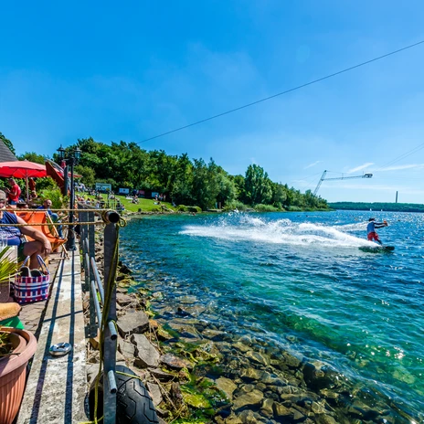 Osoba jeździ na wakeboardzie po krystalicznie czystej wodzie jeziora Kulkwitz, podczas gdy goście relaksują się na słonecznym tarasie.