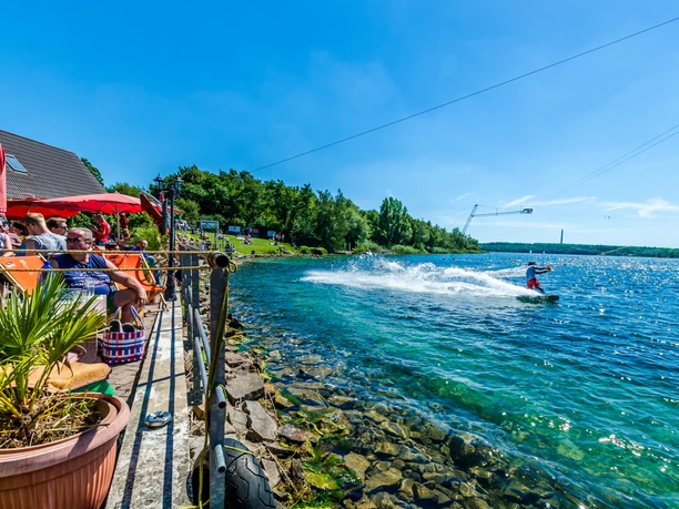 Na křišťálově čisté vodě jezera Kulkwitz jezdí člověk na wakeboardu, zatímco návštěvníci odpočívají na slunné terase.