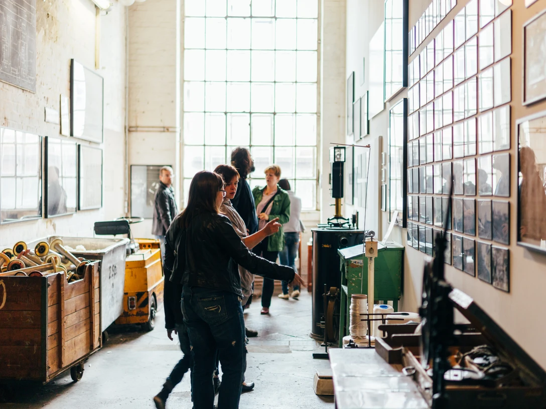 Rundgang durch die Spinnerei - Industriekultur in Leipzig Besucher besichtigen beim Rundgang durch die Alte Baumwollspinnerei eine Galerie mit zahlreichen Ausstellungsstücken und Bildern an der Wand, im Hintergrund ist ein großes Fenster zu sehen, Industriekultur, Kunst