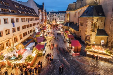 Blick von oben auf den Weihnachtsmarkt auf dem Nikolaikirchhof mit der Alten Nikolaischule auf der linken und der Nikolaikirche auf der rechten Seite, dazwischen Weihnachtsmarktbuden und einige Besucher zur Abenddämmerung
