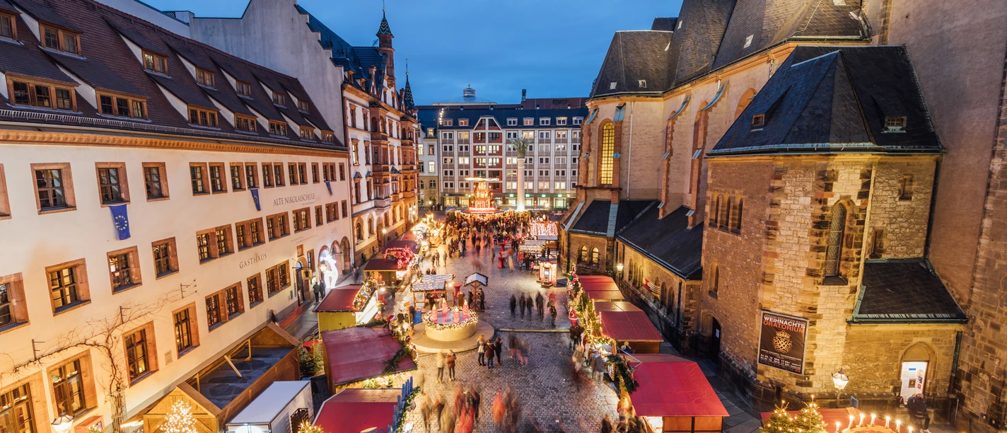 Blick von oben auf den Weihnachtsmarkt auf dem Nikolaikirchhof mit der Alten Nikolaischule auf der linken und der Nikolaikirche auf der rechten Seite, dazwischen Weihnachtsmarktbuden und einige Besucher zur Abenddämmerung