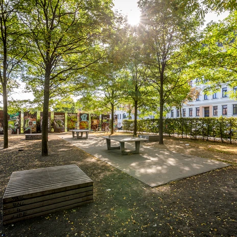 Blick auf die von Bäumen umgebenen und schattigen Tischtennisplatten im Lene-Voigt-Park; im Hintergrund: Häuserfassaden und Begrenzungen mit Graffiti