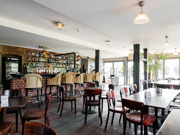 Fotografie des Innenraums des Restaurants, mit dunklem Holzmobiliar, der Bar im Hintergrund, Beleuchtung und Blick auf die großen Fenster, die helles Licht in den Saal fallen lassen, veranstaltungen, gastronomie