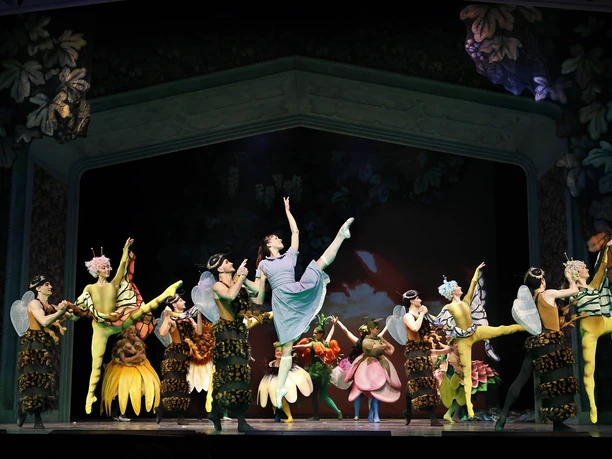 La troupe Alice au pays des merveilles se produit en costumes colorés sur une scène sombre de la Comédie musicale.