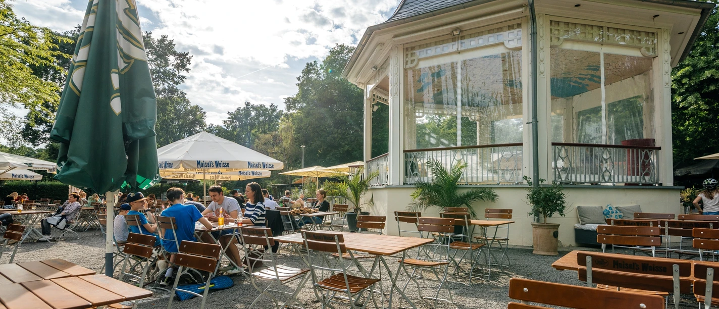 Kiosque à musique - Gastronomie à Leipzig Photographie de la terrasse et du beau pavillon blanc un jour d'été dans le parc Clara Zetkin ; avec des bois en arrière-plan, des parasols
