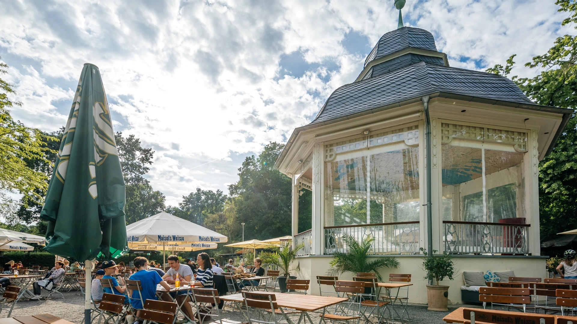 Photograph of the outdoor seating area and beautiful white pavilion on a summer's day in Clara Zetkin Park; with woodland in the background, parasols