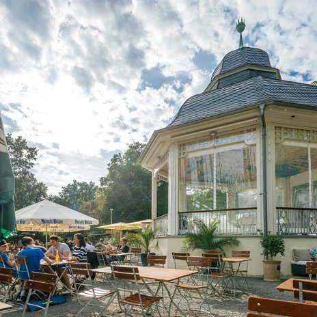Photograph of the outdoor seating area and beautiful white pavilion on a summer's day in Clara Zetkin Park; with woodland in the background, parasols