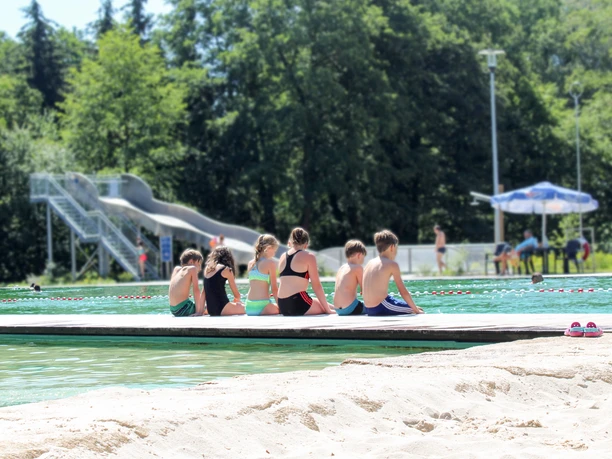 Six children sit at the edge of the pool with their backs to the camera in bathing suits and dangle their legs in the water, a large slide can be seen in the background, leisure, fun