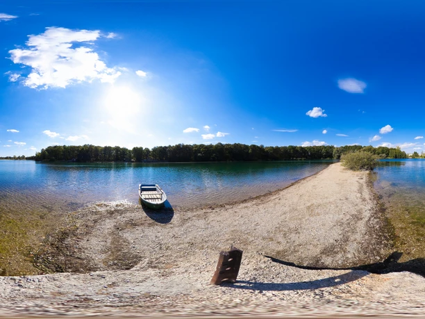 Albrechtshainer See im Leipziger Neuseenland Das Bild zeigt einen ruhigen See mit klarem Wasser, das sanft ans Ufer schwappt. Am linken Bildrand liegt ein bescheidenes Boot, bereit zur Abfahrt. Die Szenerie wird von einem strahlend blauen Himmel mit wenigen weißen Wolken überdeckt.