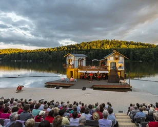 Photo de la Seebühne Kriebstein sur le barrage de Kriebstein au coucher du soleil, sur la scène est monté un décor avec 2 maisons et au milieu joue un orchestre, manifestation