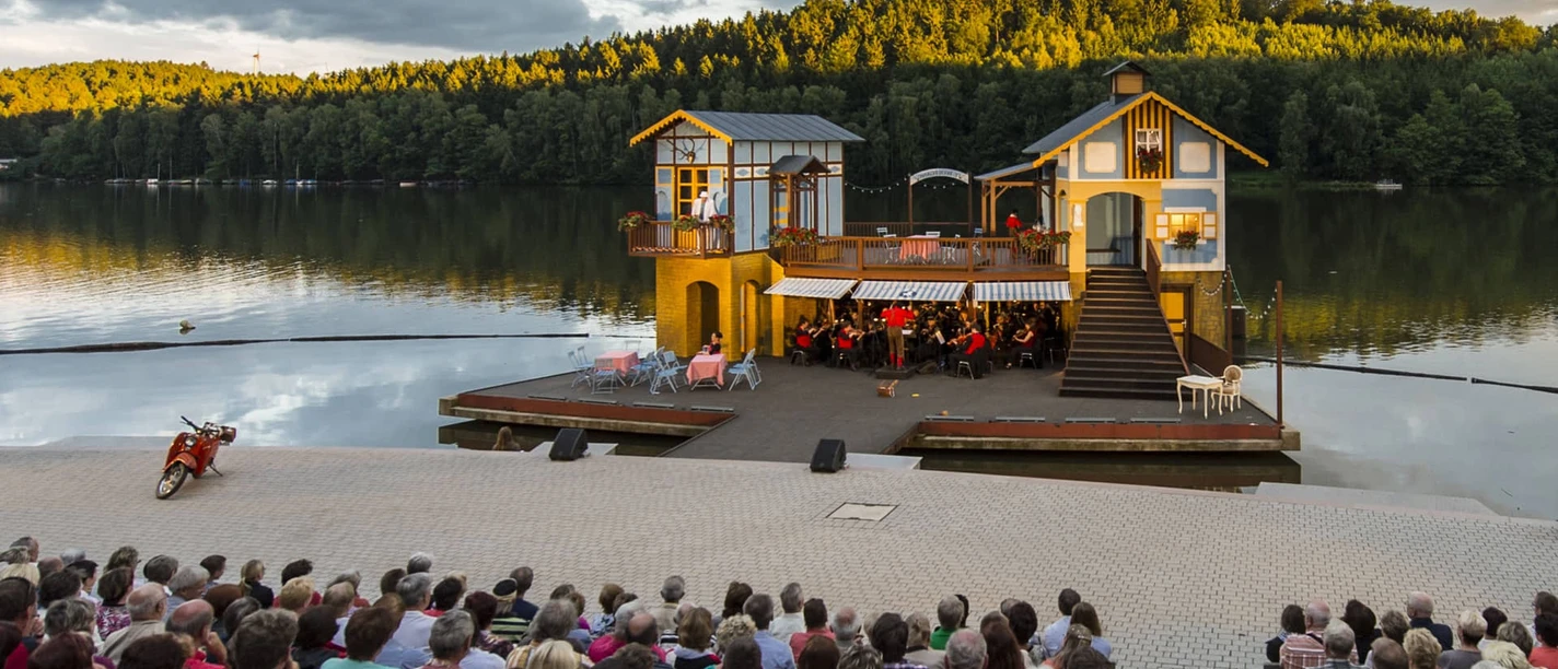 Seebühne Kriebstein - Kultur in Leipzig und Region Foto von der Seebühne Kriebstein auf der Talsperre Kriebstein bei Sonnenuntergang, auf der Bühne ist eine Kulisse mit 2 Häusern aufgebaut und in der Mitte spielt ein Orchester, Veranstaltung