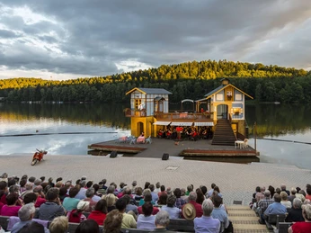 Seebühne Kriebstein - Kultur in Leipzig und Region Foto von der Seebühne Kriebstein auf der Talsperre Kriebstein bei Sonnenuntergang, auf der Bühne ist eine Kulisse mit 2 Häusern aufgebaut und in der Mitte spielt ein Orchester, Veranstaltung