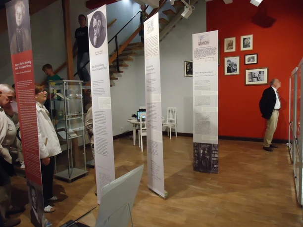 Městské muzeum Döbeln Pohled do interiéru městského muzea v Döbelnu se stojícími postavami a informačními stélami, galerie s fotografiemi na červené stěně.