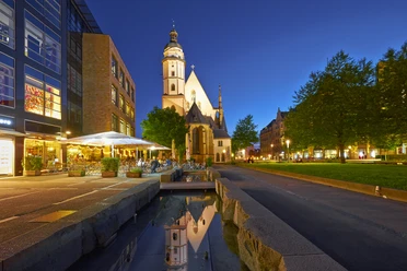 Vue sur l'église Saint-Thomas illuminée au crépuscule, à gauche devant la brasserie avec terrasse et à droite la pelouse verte de Saint-Thomas avec des arbres, une institution culturelle, des loisirs, de la musique, un ruisseau.