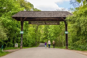 View of the entrance to Leipzig Wildlife Park in southern Connewitz in the green forest with a large wooden arch and the inscription "Wildpark Leipzig", which a family is just passing through