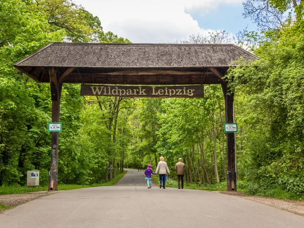 Pohled na vchod do lipského přírodního parku v jižním Connewitzu v zeleném lese s velkým dřevěným obloukem a nápisem "Wildpark Leipzig", kterým právě prochází rodina.