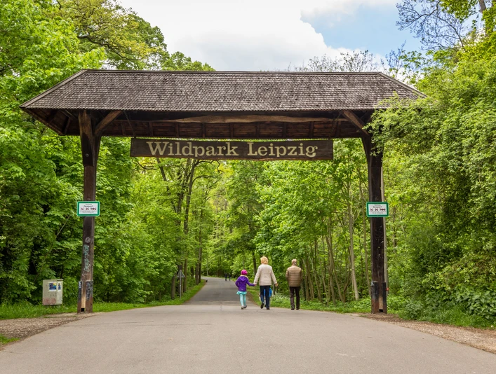 Wildpark Leipzig - Tierisches Leipzig Blick auf den Zugang zum Wildpark Leipzig im südlichen Connewitz im grünen Wald mit einem großen Holzbogen und der Aufschrift "Wildpark Leipzig", den gerade eine Familie passiert