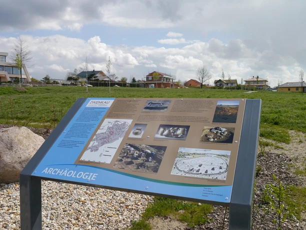 Un panneau d'information au bord du lac Zwenkauer montre des objets archéologiques dans un paysage rural.