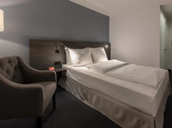 Zimmer Queen 06_Lindgart-Hotel-Z2_030-HDR.jpeg Modernes Hotelzimmer mit elegantem Doppelbett, einladendem Sessel und beruhigendem Farbton.