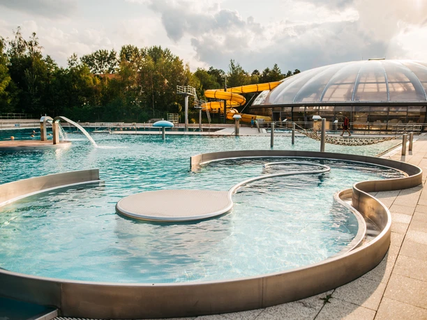 Johannisbad Außenthermalbad mit Wasserrutsche, umgeben von Bäumen, unter wolkigem Himmel.