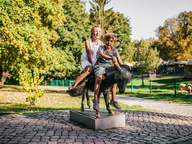 Two laughing children on a goat statue in the zoo, surrounded by trees.