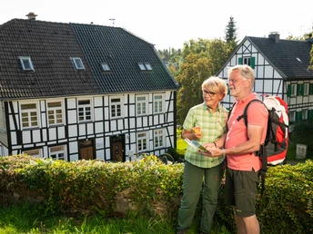 Unterwegs in Haan-Gruiten Ein älteres Paar mit Freizeitkleidung und Wanderkarte steht vor Fachwerkhäusern in einer sonnigen Umgebung.