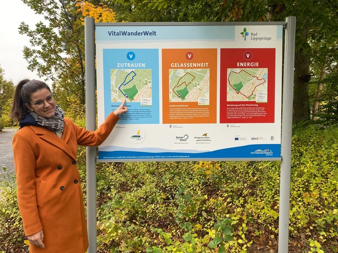 Eine Person im orangefarbenen Mantel zeigt auf ein Informationsschild im Herbstwald. Schilder beschreiben Wanderwege.