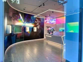2021_Product Exhibition_Experience More Light(3).jpg Ausstellungsbereich der JENOPTIK AG mit beleuchteten Vitrinen, Produkten und einem bunten Wandgemälde mit dem abstrahierten Portrait von Carl Zeiss