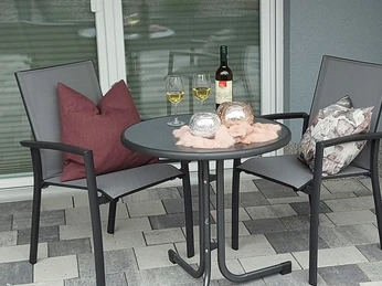 Terrasse einer modernen Ferienwohnung mit zwei Stühlen, Tisch und Weinflasche vor einer Glastür.
