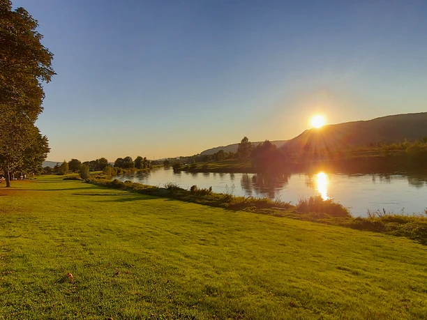 moderne_ferienwohnung_Weser.jpg Sonnenaufgang über der Weser mit grünem Ufer und Bäumen, ruhiges Wasser, klare Morgenstimmung.