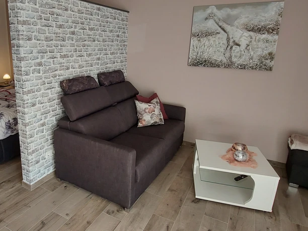 Wohnzimmer mit grauem Sofa, Couchtisch, Gemälde an der Wand, Bett im Hintergrund und Holzboden.