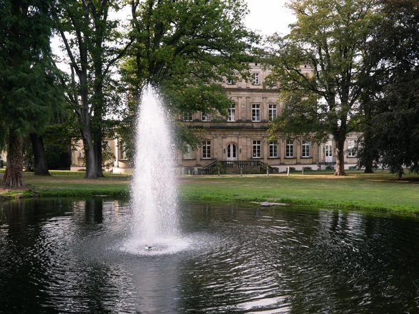 Teutoburger_Wald_Detmold_B.Fromberger_Palaisgarten_CCBYSA (17).jpg Ein Springbrunnen im Vordergrund eines historischen Gebäudes in einem grünen Park mit Bäumen.