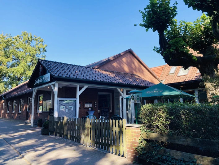 Eingang Restraurant Restaurantfront mit roten Ziegeln, weißen Holzleisten und einem Garten mit Sonnenschirm.