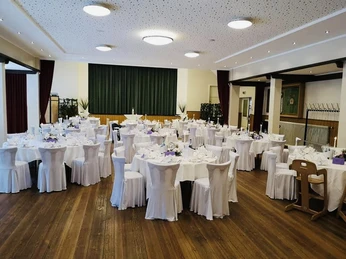 Saal Festlich gedeckter Veranstaltungssaal mit runden Tischen, weißen Stuhlhussen und gedimmter Beleuchtung.