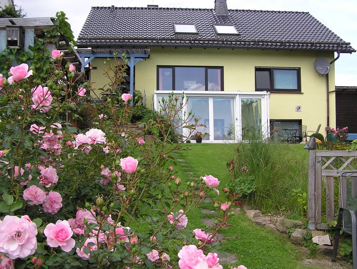 Hausansicht Blick aus dem Garten: Hausansicht mit Wintergarten, gepflegtem Garten und Rosenbüschen im Vordergrund.