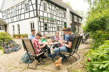 Pause im Hofcafé auf dem Hof zur Hellen im Windrather Tal Menschen sitzen an einem Tisch im Freien eines Fachwerkhauses. Ein Huhn spaziert um den Tisch.
