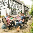 Pause im Hofcafé auf dem Hof zur Hellen im Windrather Tal Menschen sitzen an einem Tisch im Freien eines Fachwerkhauses. Ein Huhn spaziert um den Tisch.