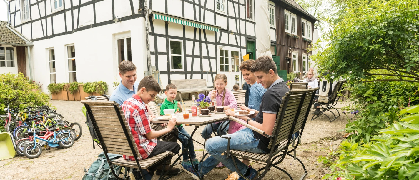 Pause im Hofcafé auf dem Hof zur Hellen im Windrather Tal Menschen sitzen an einem Tisch im Freien eines Fachwerkhauses. Ein Huhn spaziert um den Tisch.