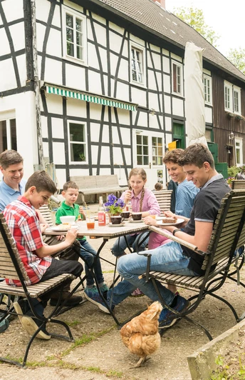 Pause im Hofcafé auf dem Hof zur Hellen im Windrather Tal Menschen sitzen an einem Tisch im Freien eines Fachwerkhauses. Ein Huhn spaziert um den Tisch.