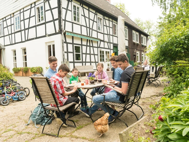 Pause im Hofcafé auf dem Hof zur Hellen im Windrather Tal Menschen sitzen an einem Tisch im Freien eines Fachwerkhauses. Ein Huhn spaziert um den Tisch.