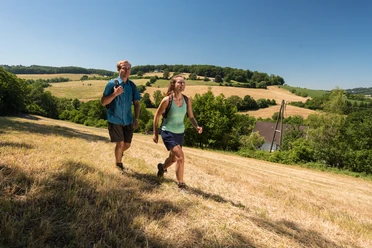 Wandergenuss in der Elfringhauser Schweiz Zwei Personen wandern auf einem grasbewachsenen Hügel mit weiten Feldern und Bäumen im Hintergrund.