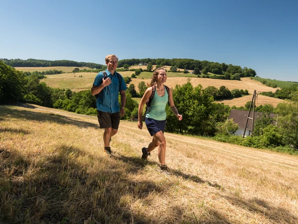 Wandergenuss in der Elfringhauser Schweiz Zwei Personen wandern auf einem grasbewachsenen Hügel mit weiten Feldern und Bäumen im Hintergrund.