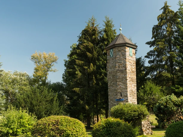 Blick auf den Turm von Schloss Hetterscheidt bei der Abtsküche in Heiligenhaus Ein steinerner Turm umgeben von hohen Bäumen und grünen Büschen, unter einem blauen Himmel.