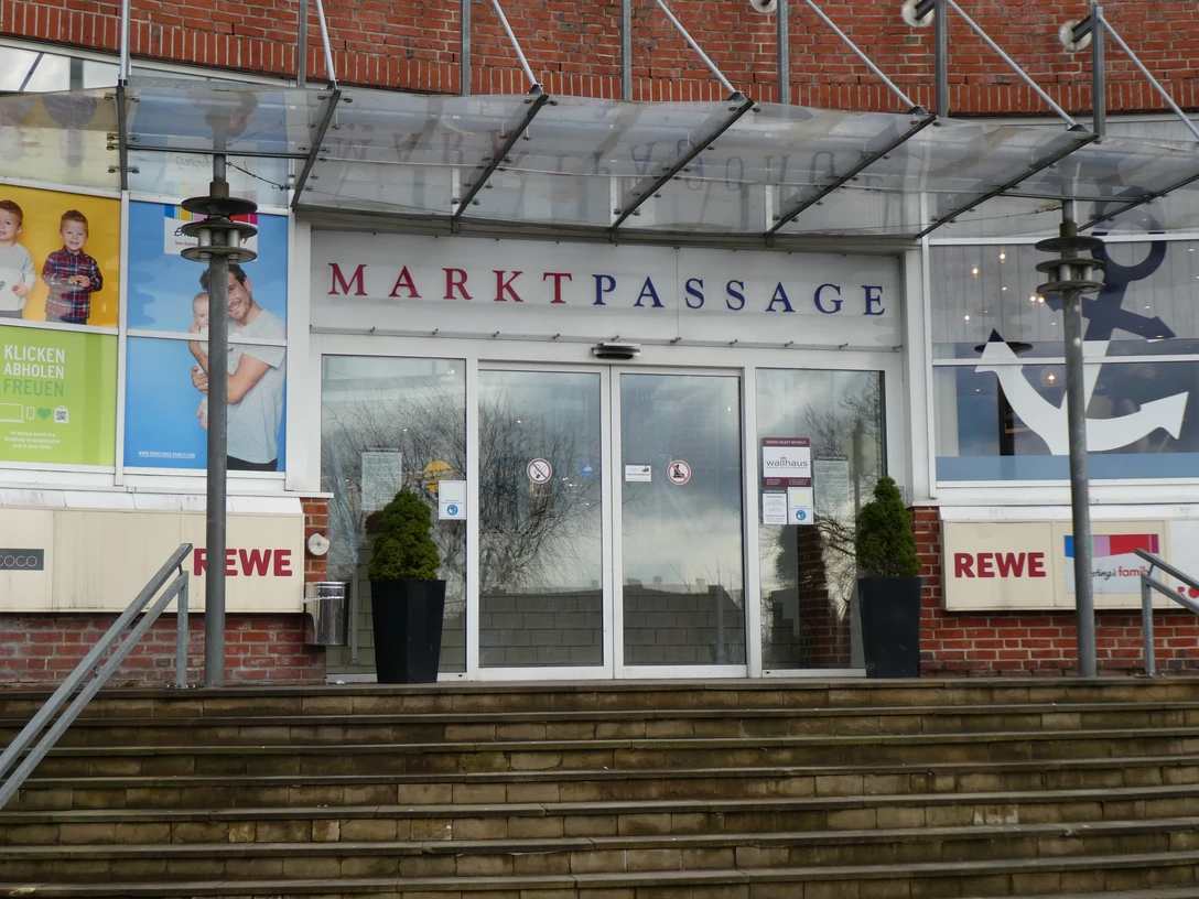 Eingang zur Markpassage mit Glastüren, flankiert von zwei Pflanzkübeln auf einer steinernen Treppe.