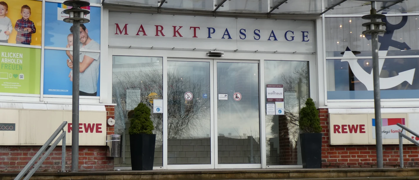Eingang zur Markpassage mit Glastüren, flankiert von zwei Pflanzkübeln auf einer steinernen Treppe.