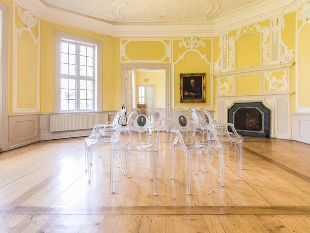 Gartensaal des Lessinghauses Heller Raum mit gelben Wänden mit Stuckverzierungen, in der Mitte transparente Stühle mit abgebildeten Personen