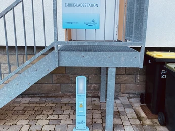 IMG_7511.jpeg E-Bike-Ladestation unter einer Metalltreppe an einer Hauswand mit Pflastersteinen davor.
