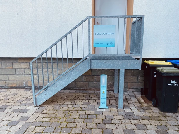 Treppe mit Handlauf vor Gebäude, daneben steht eine E-Bike-Ladestation auf gepflastertem Boden.